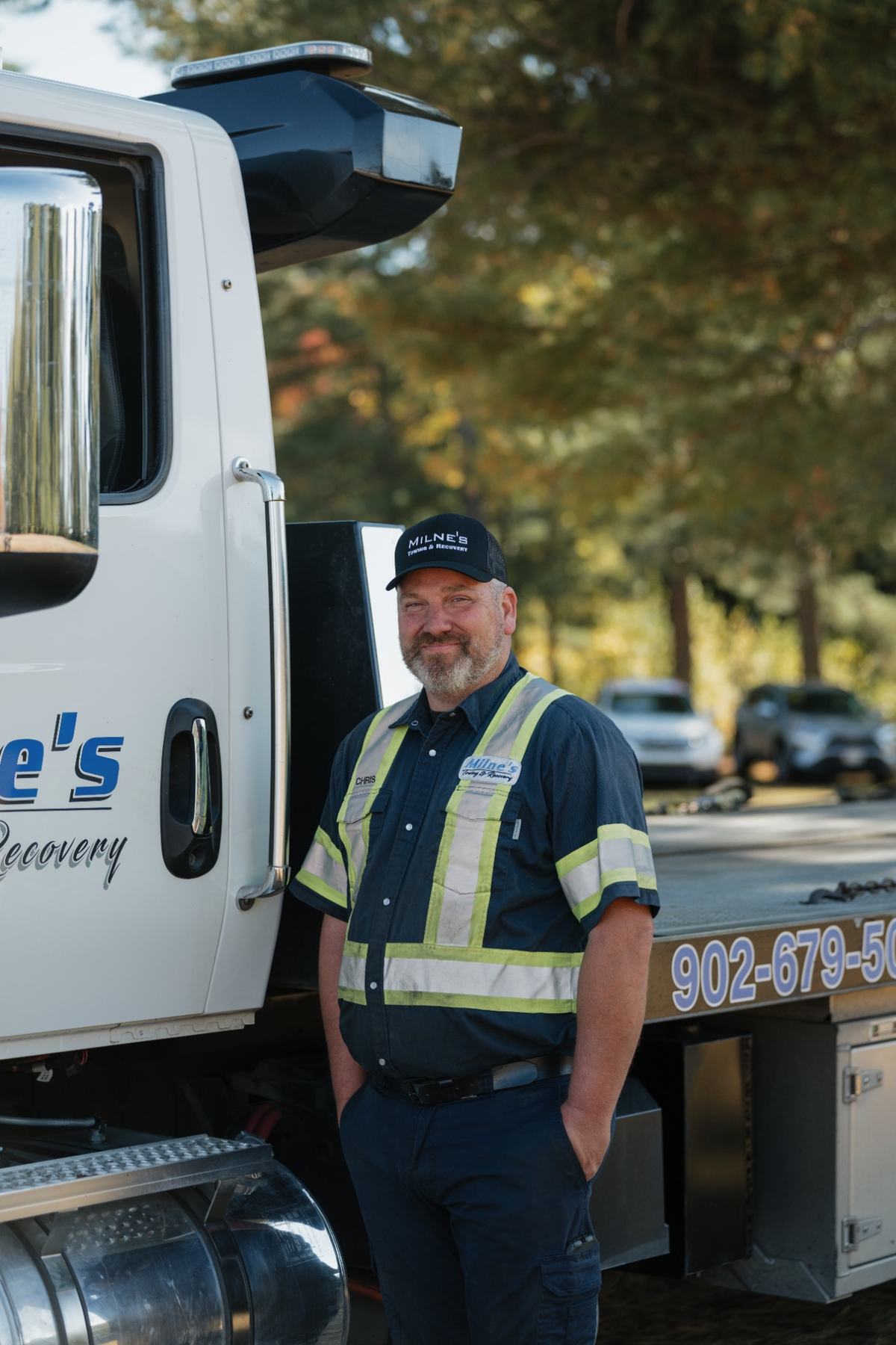 Chris, in a Milne's cap and navy work shirt with reflective stripes, standing beside a Milne's flatbed.