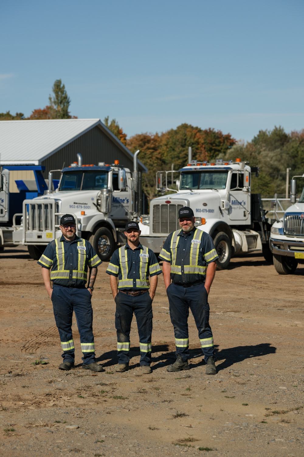 Rob, Jason and Chris standing side by side in the yard with two white Milne's tow trucks behind them.
