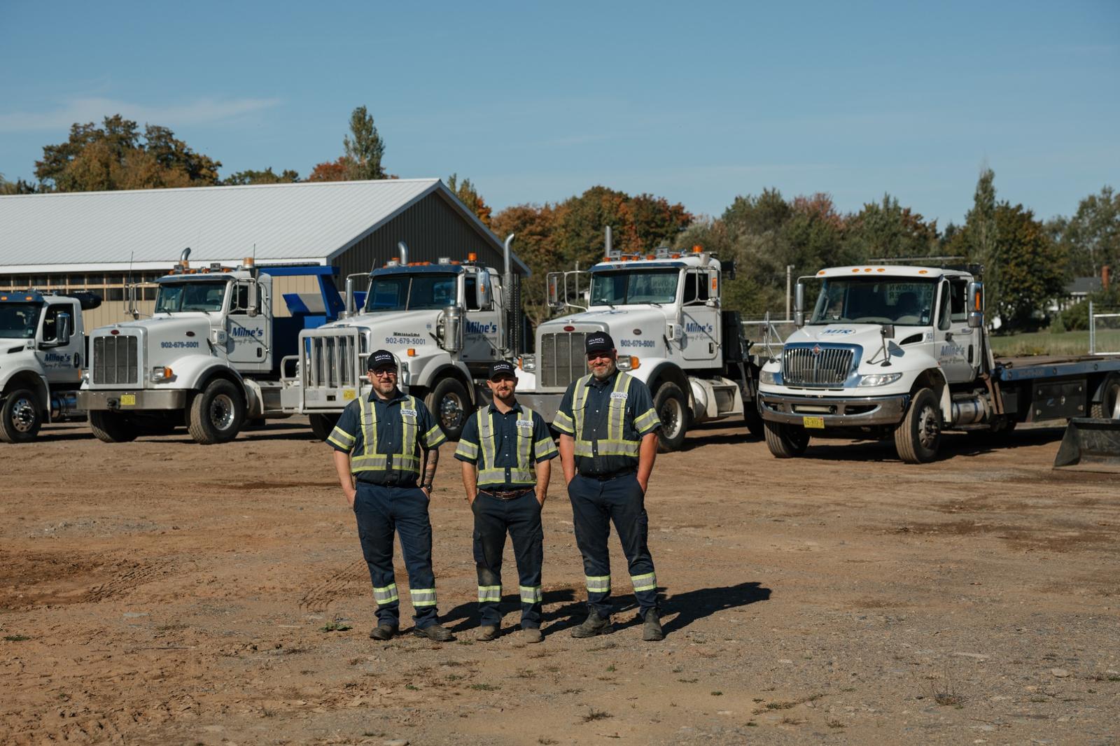 Rob, Jason and Chris in navy work shirts with reflective stripes, standing in the yard in front of a row of white Milne's tow trucks.