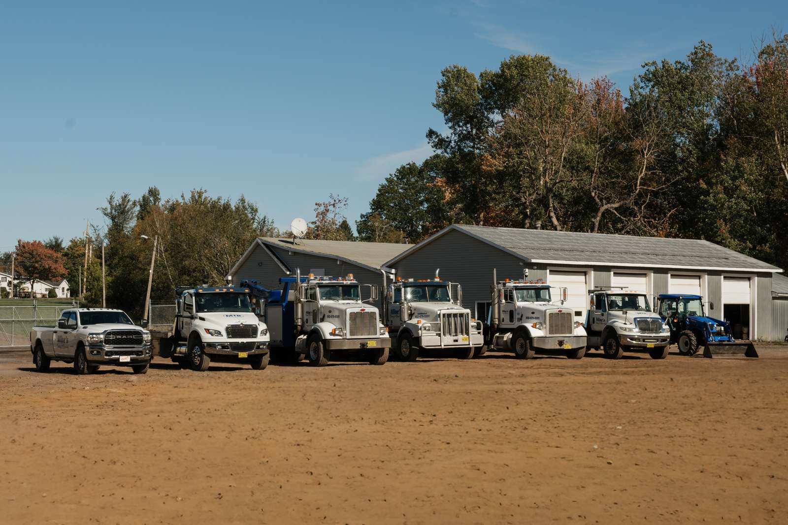 Now: the Milne's fleet today, a pickup, a row of white heavy tow trucks and wreckers and a blue loader tractor, lined up in front of a grey four-bay shop.