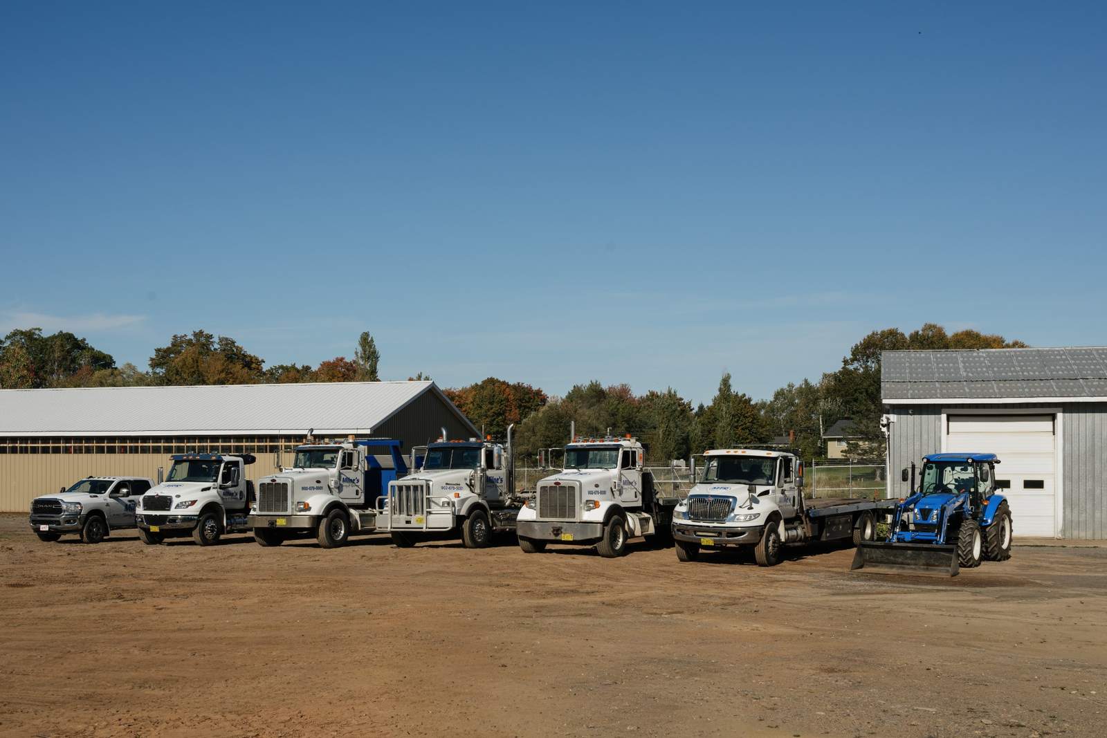 The Milne's fleet parked in a row on the gravel yard in front of the shop on Highway 359, under a clear blue sky.