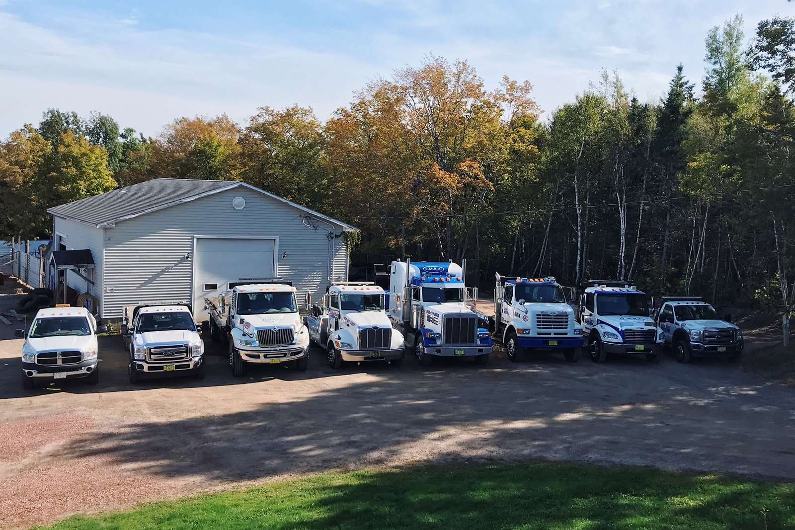 Then: the earlier Milne's fleet, eight white and blue pickups and tow trucks, lined up on a gravel lot beside a grey single-bay garage under autumn trees.