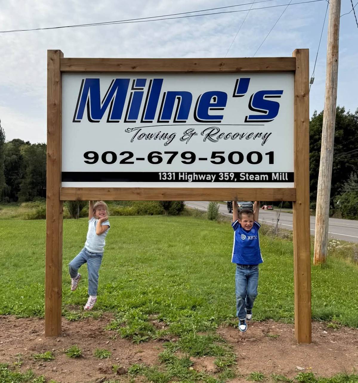 Brenna and Rob’s two children hanging from the bottom rail of the new Milne’s Towing & Recovery sign at 1331 Highway 359, Steam Mill.