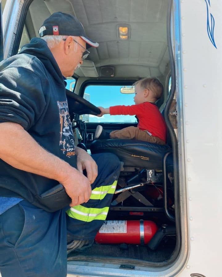 Lindsay Milne at the open door of a truck cab while his young grandchild, in a red sweater, sits in the driver’s seat holding the wheel.
