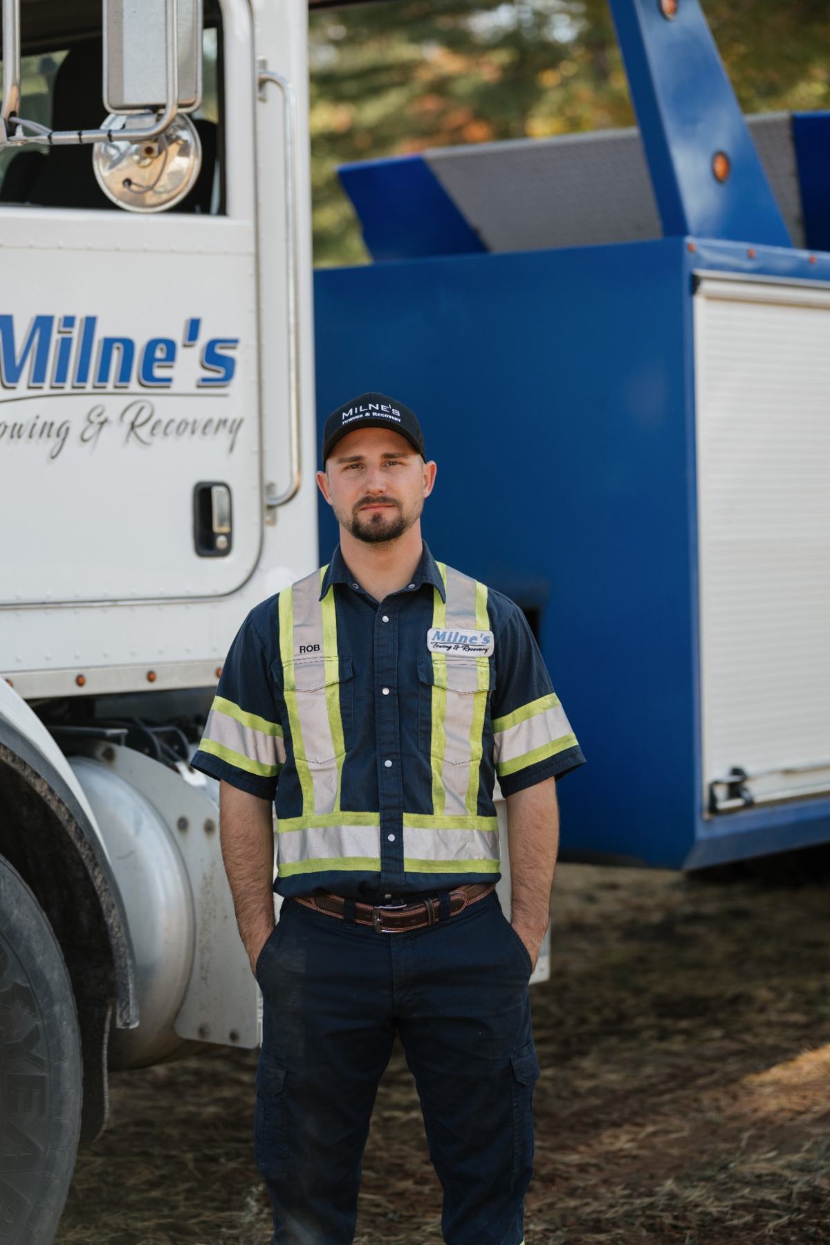 Rob, in a Milne's cap and navy work shirt with reflective stripes, standing in front of the blue heavy wrecker.