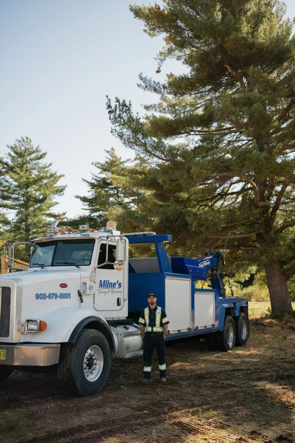 Milne's blue and white heavy wrecker, unit 01, parked under tall pines with a crew member standing beside it.